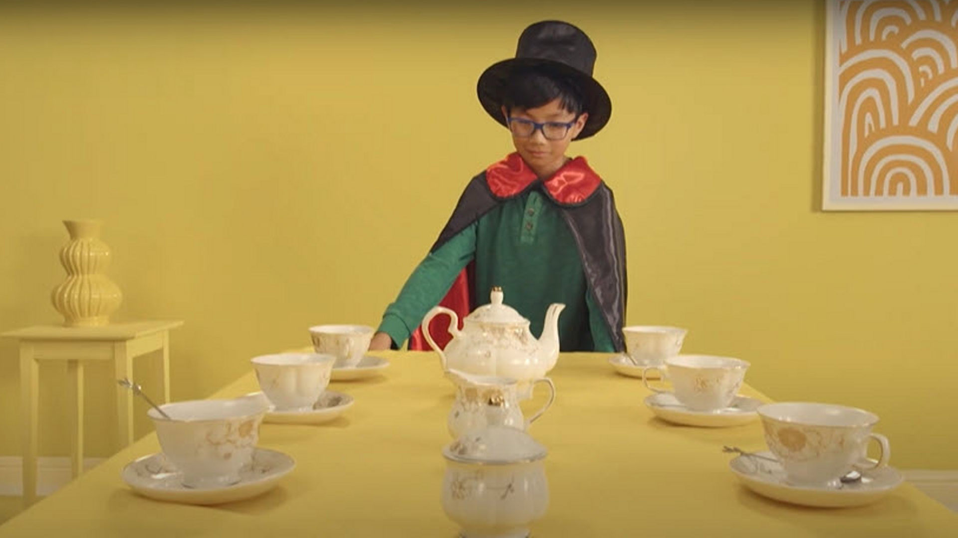 Young child wearing cape and hat at dining table.